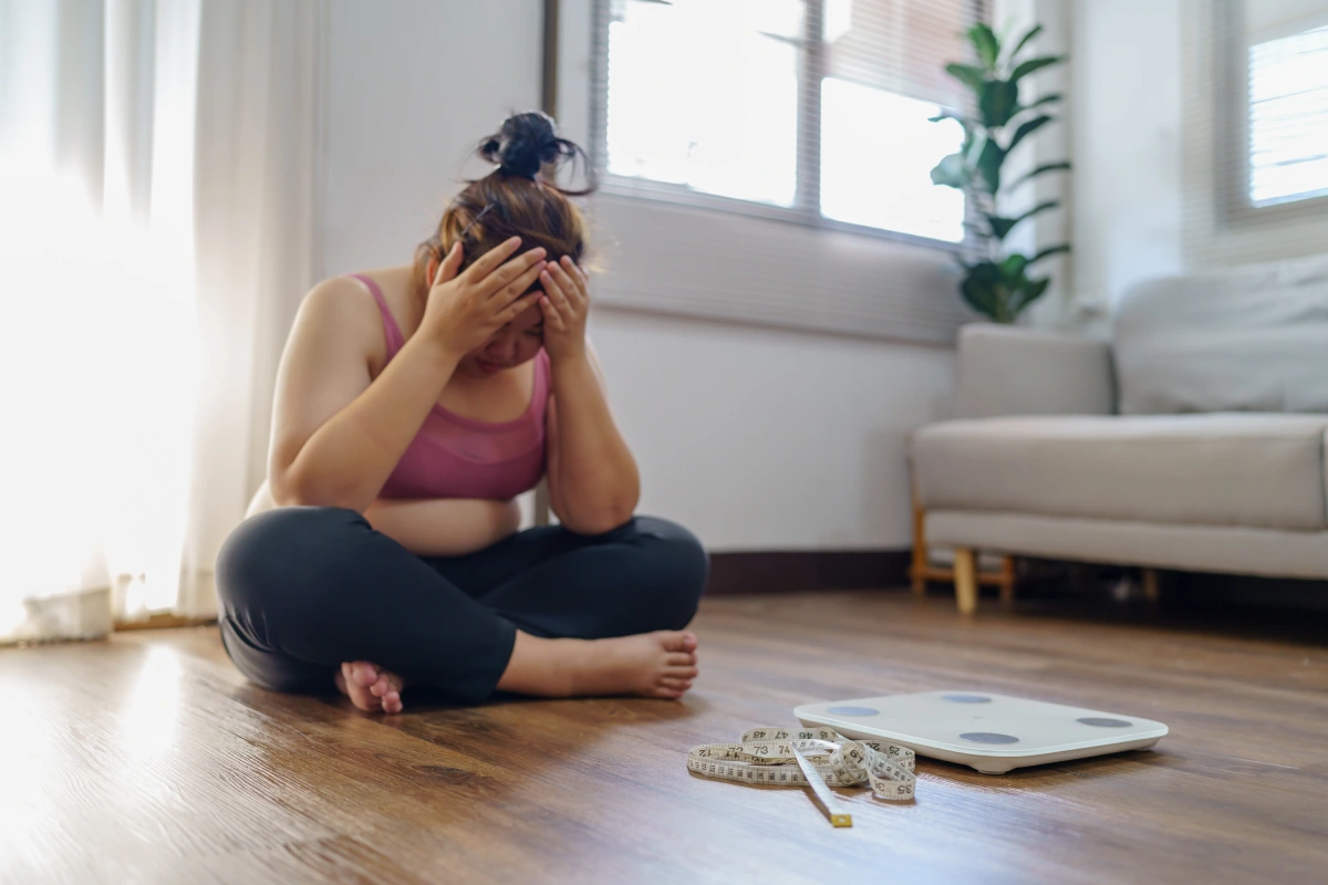 A frustrated woman sitting on the floor looking at a weight scale, highlighting the struggles of weight gain addressed by peptide therapy at Novu Wellness and Aesthetics.